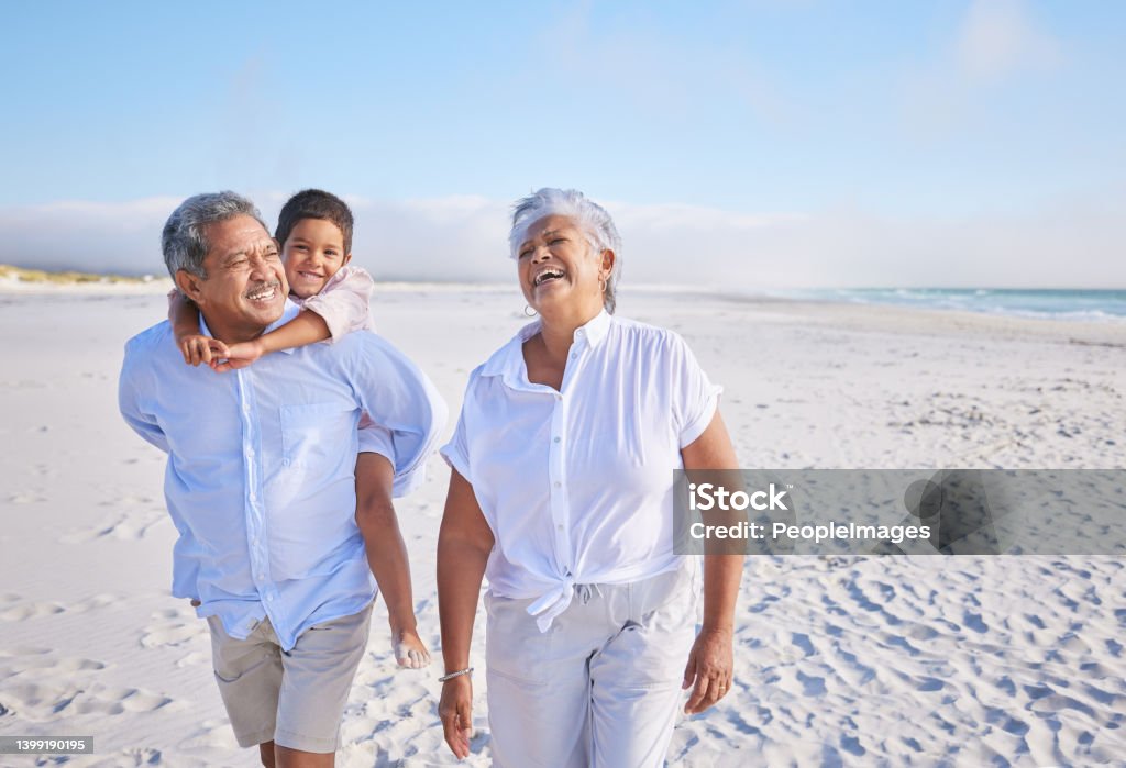 Happy mixed race grandparents walking on the beach with their grandson. Little boy enjoying a piggyback ride on his grandfathers back during summer vacation by the beach Senior Adult Stock Photo