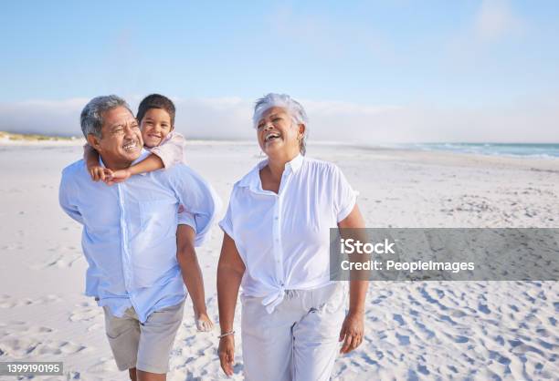 Happy Mixed Race Grandparents Walking On The Beach With Their Grandson Little Boy Enjoying A Piggyback Ride On His Grandfathers Back During Summer Vacation By The Beach Stock Photo - Download Image Now