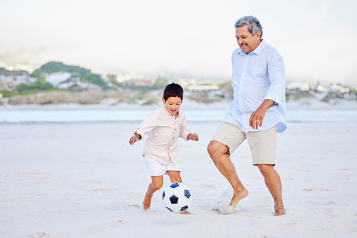 Active grandfather and grandson playing soccer on sandy beach. Cute little boy kicking ball with his grandpa