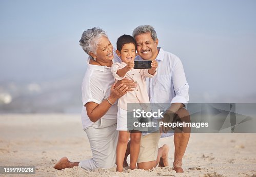istock Grandparents with grandson at the beach holding mobile and taking selfie or doing video call with family during vacation by the sea. Adorable little boy taking a picture with his grandmother and grandfather 1399190218