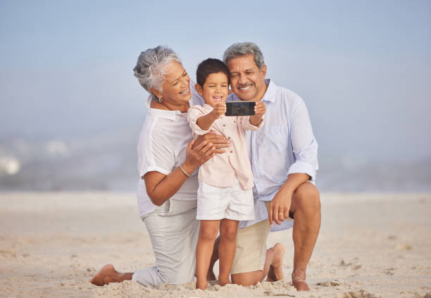 grandparents with grandson at the beach holding mobile and taking selfie or doing video call with family during vacation by the sea. adorable little boy taking a picture with his grandmother and grandfather - grandparent retirement senior adult healthy lifestyle imagens e fotografias de stock