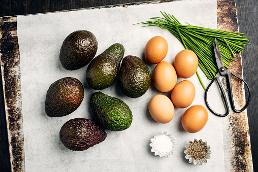 Overhead view of the ingredients for a dish of baked eggs with avocado. Recipe, cut the avocado in half and remove the stone, use small sized eggs, discard some of the egg white if desired and pour the remaining yolk and white into the hole left by the avocado stone, season with salt and pepper and some chives and or some chilli flakes. Bake for around 10-12 minutes at 200 degrees until the egg whites have set but the yolks are still runny. Serve with toasted bread, delicious.