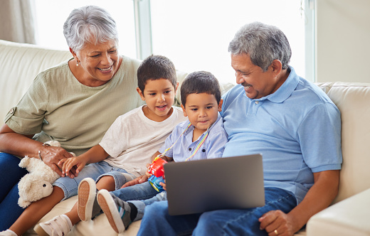 Mixed race senior couple bonding with their grandsons and using a laptop on the sofa at home. Hispanic senior man and woman having fun and smiling with their grandkids