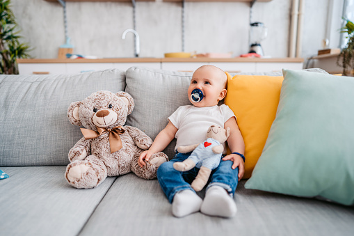 Close-up of a newborn baby boy sitting down on the sofa with a teddy bear next to him.