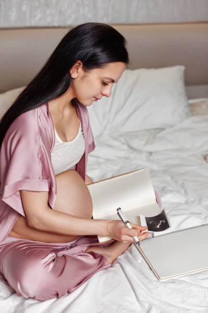 istock Pregnant woman sitting on bed and writing in notebook 1399198428