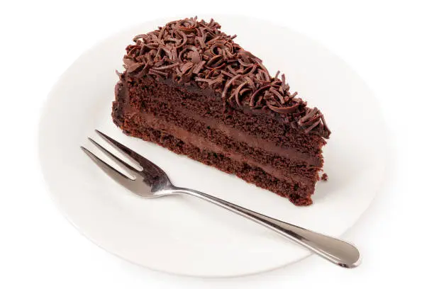 Slice of chocolate cake with cream filling and chocolate shavings next to fork on white plate isolated on white.