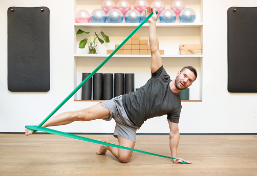 Full body of barefoot male athlete doing side kick exercise with elastic band while training on floor in modern fitness studio. Man fitness concept.