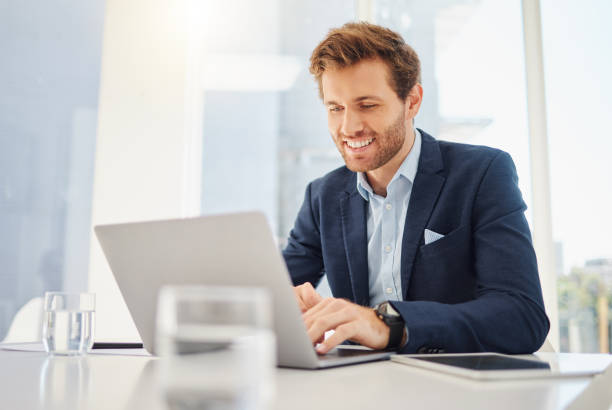 un jeune homme d’affaires caucasien heureux travaillant sur un ordinateur portable dans un bureau. entrepreneur masculin confiant planifiant tout en naviguant sur internet et en envoyant des courriels aux clients. homme rejoignant une conférence virtuel - planning business person clothing well dressed photos et images de collection