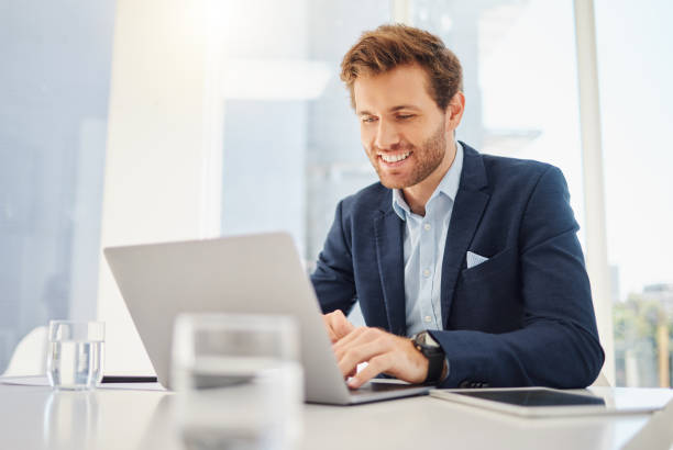 one happy young caucasian businessman working on a laptop in an office. confident male entrepreneur planning while browsing the internet and sending emails to clients. man joining a virtual conference via webcam - lawyer young adult suit expressing positivity imagens e fotografias de stock