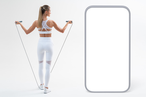 Back view of fit blonde woman dressed in crop top, leggings and sketchers doing cardio workout with skipping rope ,with big mockup phone on the left with empty screen for promotional content