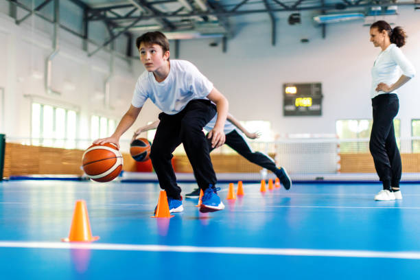 Physical education class and sport training in high school Students and their coach are in school gym and have training class. Teacher give instructions to pupils and watch them with attention. Boys are running in precise order between the cones with basketball. They are wearing sports uniform and warming up. youth basketball indoor stock pictures, royalty-free photos & images