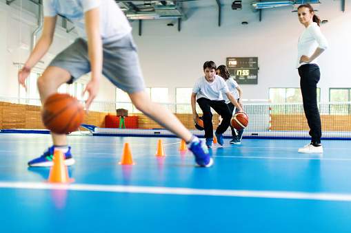 Students and their coach are in school gym and have training class. Teacher give instructions to pupils and watch them with attention. Boys are running in precise order between the cones with basketball. They are wearing sports uniform and warming up.