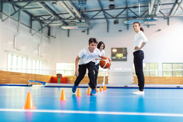 Students and their coach are in school gym and have training class. Teacher give instructions to pupils and watch them with attention. Boys are running in precise order between the cones with basketball. They are wearing sports uniform and warming up.