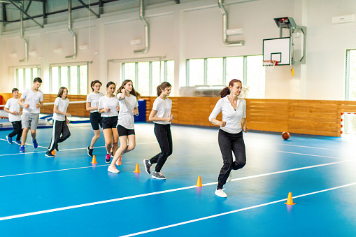Students and their coach are in school gym and have training class. Teacher give instructions to pupils and she is on front of them. Boys and girls are running in precise order between the cones. They are wearing sports uniform and warming up.