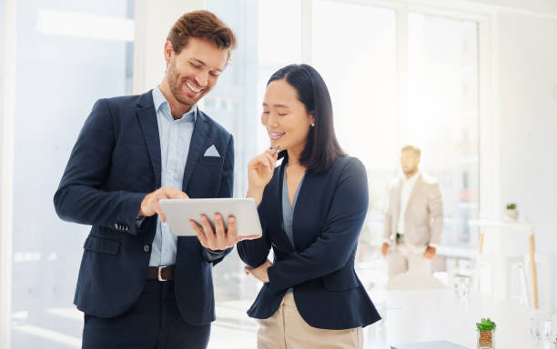 deux jeunes collègues heureux et divers discutent ensemble de plans et d’idées sur une tablette numérique dans un bureau. une femme d’affaires asiatique et un homme d’affaires caucasien réfléchissent en ligne avec des applications intelligentes - cheerful business person strategy people photos et images de collection