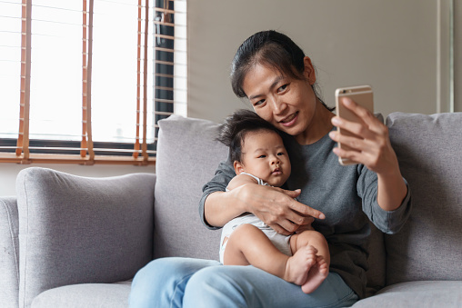 Lovely young Asian mother and newborn baby boy having video call via mobile phone together. Mom using smartphone and taking selfie with her little cute baby while sitting on sofa at home.