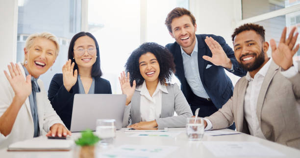 portrait of diverse corporate businesspeople waving hello to colleagues during a virtual teleconference meeting via video call in an office boardroom. happy staff greeting during online global webinar - welcome back imagens e fotografias de stock