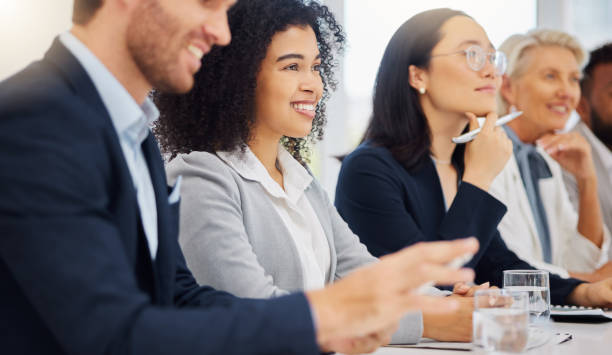 feliz jovem empresária mestiça ouvindo uma apresentação durante uma reunião em uma sala de reuniões de escritório ao lado de seus colegas. diversos grupos de empresários sorridentes sentados em uma fileira em um painel como público durante um work - panel dicussion - fotografias e filmes do acervo