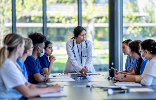 Medical Residence Team Meeting A small group of medical student residents gather around a boardroom table to to meet with their medical team lead.  They each have cases out on front of them as they work together collaboratively to discuss each one.  They are each dressed professionally in medical scrubs and are listening attentively to the doctor leading the meeting at the head of the table. medical student stock pictures, royalty-free photos & images