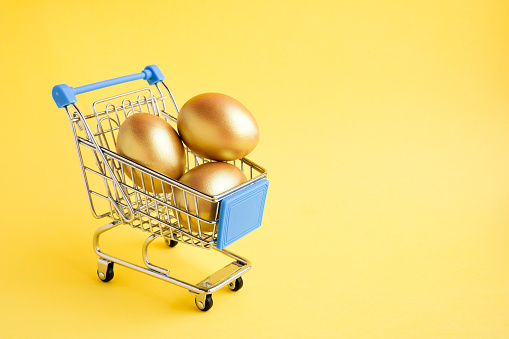 Shopping cart with three golden eggs isolated on yellow background. Easter shopping or sale banner with copy space for text. Concept of a good buy, successful investment, making money.