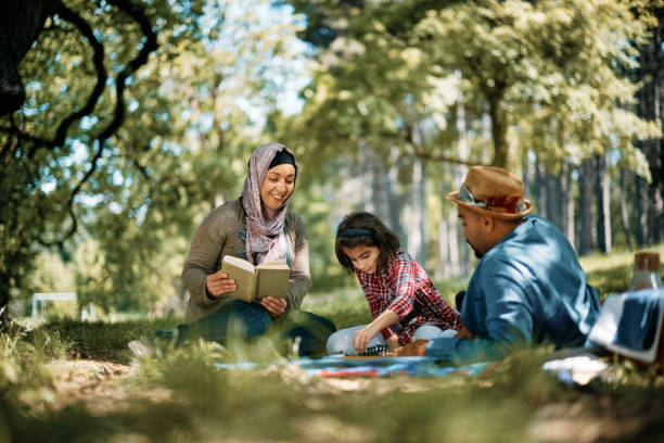 Happy Muslim parents and their daughter having family picnic in springtime. Happy Muslim woman reading book while her husband and daughter are playing chess during their picnic day in nature. Copy space. middle eastern ethnicity mature adult book reading stock pictures, royalty-free photos & images