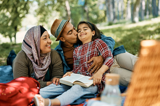 Happy Muslim parents and their daughter relaxing during family picnic in springtime. Father is kissing his daughter.