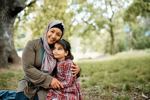 Happy Muslim woman embracing her small daughter in nature and looking at camera. Copy space.