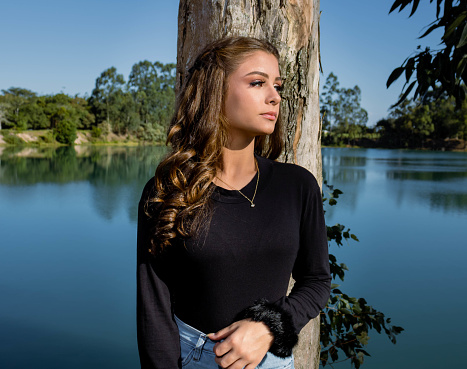 Girl with green eyes wearing blue jeans and a black blouse next to a lake of crystal clear water in green and blue tones with mountains and trees in the background. Mention the discovery of new places