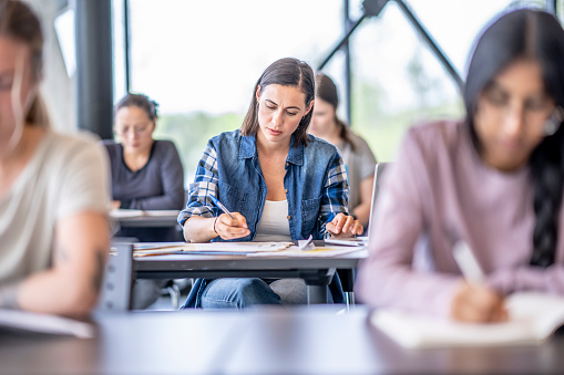 A small group of diverse University students sit in rows of desks, as they work individually on their assignments.  They are dressed casually and are focused on their work.  The focus is on a female student in the center who is working away on a writing assignment.