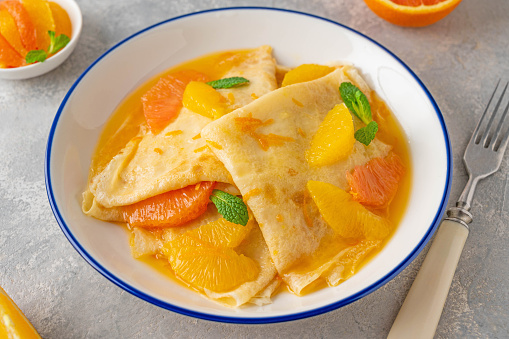 Delicious crepes suzette with orange syrup and slices fruits on a plate on a gray concrete background, top view.
