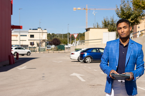A young latin black man in blue jacket standing and holding a folder posing looking at camera in a parking lot.