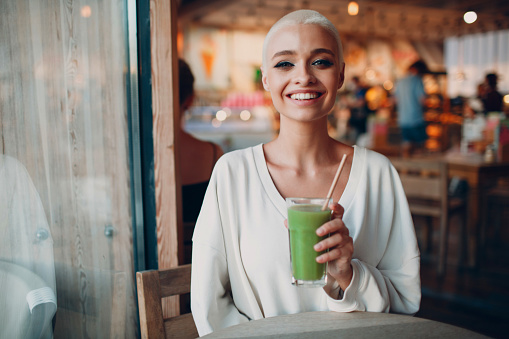 Portrait of young smiling millenial european short haired woman with green smoothie at cafe. Beautiful happy blonde girl indoor.