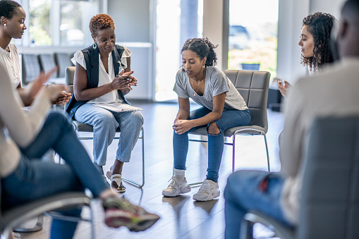 A small group of diverse adults sit together on chairs in a circle.  They are each dressed casually and are sharing their struggles with one another in a group therapy session.  A mature woman of African decent is leading the session and a young woman is sharing with the group.