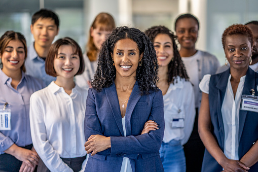 A large group of ethnically diverse business professionals pose for a portrait.  They are spaced evenly apart and are each dressed in business attire as they cross their arms and smile.  The focus is on a young woman in front of African decent.