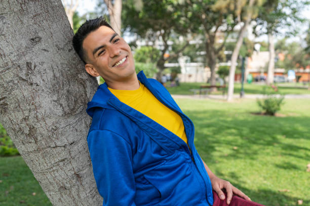 istock Latin man smiling as he leans against a tree in a park 1399285233