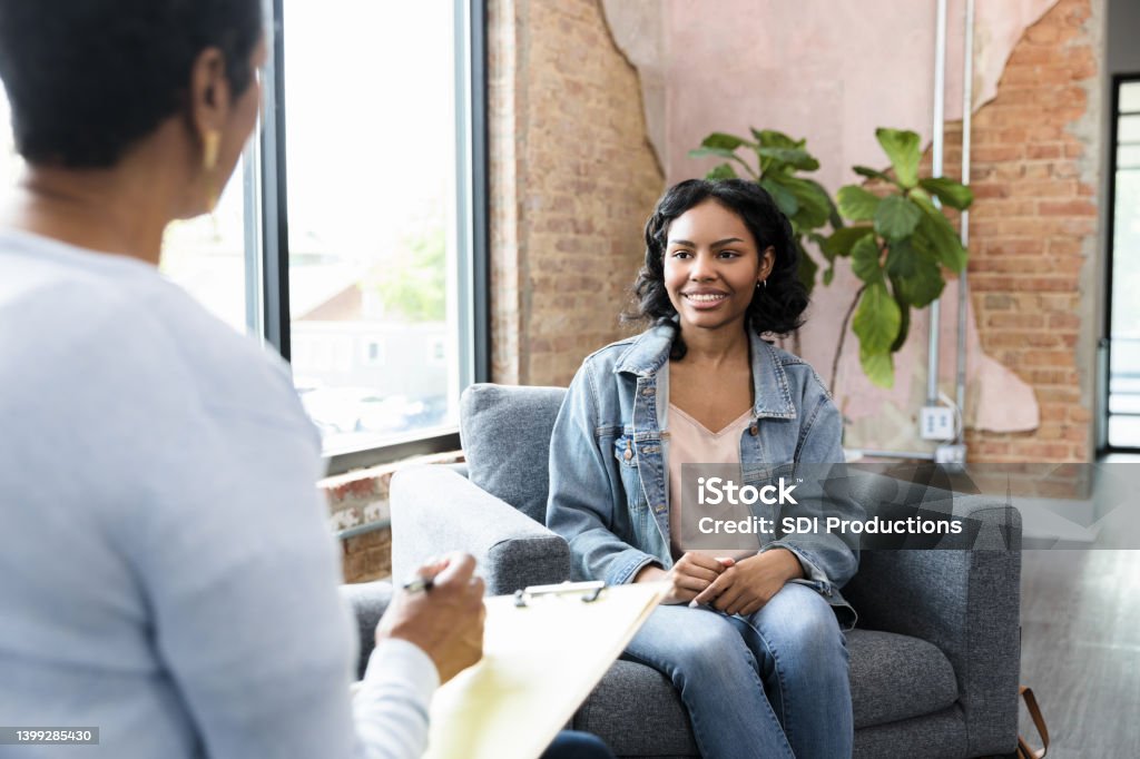 Young woman smiles while answering therapist's questions As the unrecognizable female counselor asks her questions, the young adult woman smiles and answers. Psychotherapy Stock Photo