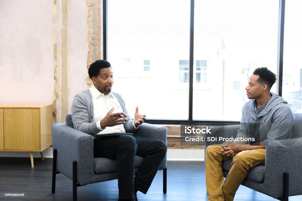 Mid adult male counselor gives young man advice When they meet together, the mid adult male therapist gestures and gives the young adult man advice. Psychotherapy Stock Photo