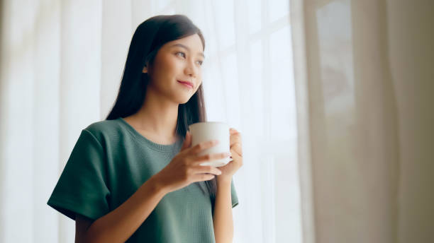 若いアジアの女性は窓のそばに立って、自宅で寝室でマグカップを保持しています, 彼女は朝起きた後にミルクを飲んでいます - time for tea ストックフォトと画像