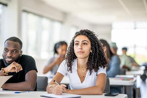 A female University student of mixed race sits at her desk surrounded by her peers as she takes notes during a lecture.  She is dressed casually and has a notebook out on the desk in front of her as she writes down points to remember.