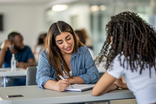 Two female College students sit at a desk as they work together on an assignment.  They are both dressed casually and have a notebook out in front of them as they jot down ideas and brainstorm together. Their peers can be seen working in small groups in the background as well.