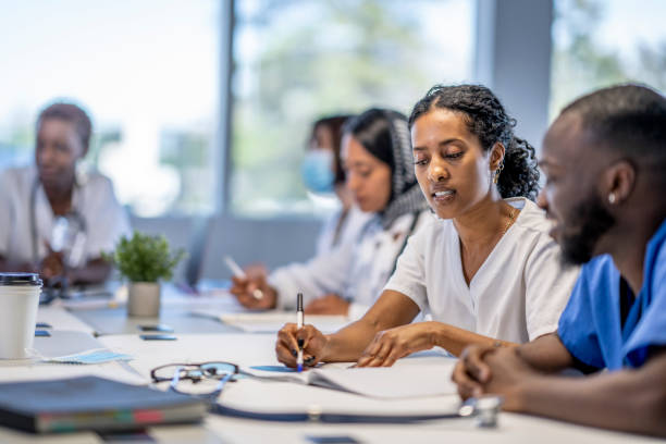 Collaborative Medical Team Meeting A small group of medical professionals sit around a boardroom table as they meet to discuss patient cases.  They are each dressed professionally in scrubs and lab coats as they work through the case files spread out on the desk collaboratively in small groups. medical student stock pictures, royalty-free photos & images