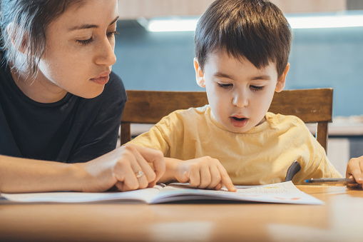 Caucasian little boy and his mother sitting together at table and reading book, mother teaching her child to read. Education, learning, study. Mother language.