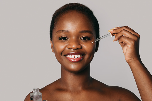Smiling nude african-american woman portrait holding opened bottle with moisturizing skin serum, pampering herself isolated over grey background. Facial skin treatment. Beautiful black woman. Facial skincare.