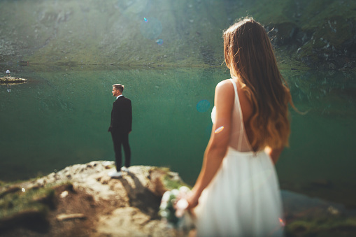 The bride approaching from behind the groom, who is waiting on the mountainside looking at the landscape with mountains and lake. Nature landscape. Wedding travel. Holiday wedding.