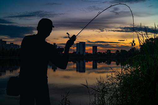 Night evening fishing at urban lake. Silhouette of fisherman with spinning rod on colorful sky. Street fishing concept