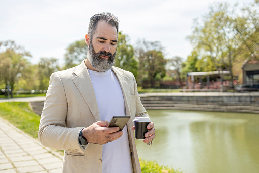 Mature man walking by the lake when using smart phone and holding cup of coffee