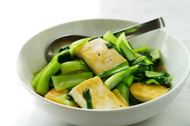 Healthy homemade vegetarian food, stir-fried Bok Choy with fried soft tofu and oyster sauce in a white bowl on a marble table. Bok Choy, Pak Choi, or Pok Choi is a type of Chinese cabbage.