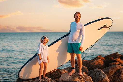 Summer activity vacations. Happy father and daughter standing with sup board on big beach rocks. Sunset sky and ocean at the background. Surfing.