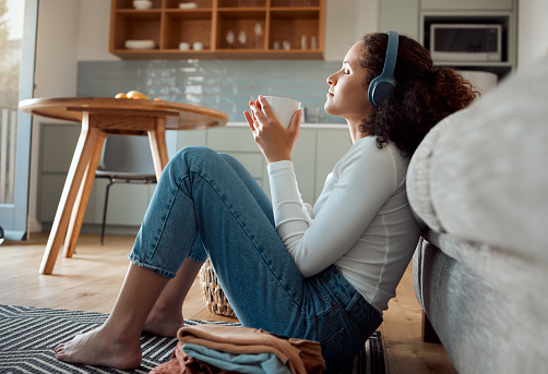 Woman enjoying a cup of tea. Young woman listening to music in her headphones drinking coffee. Hispanic woman drinking a cup of tea sitting on the floor. Woman relaxing, enjoying a beverage at home