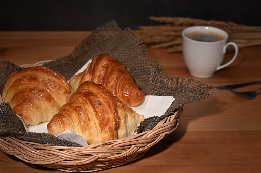 Tasty buttery croissants in wicker basket and coffee cup on wooden table.
